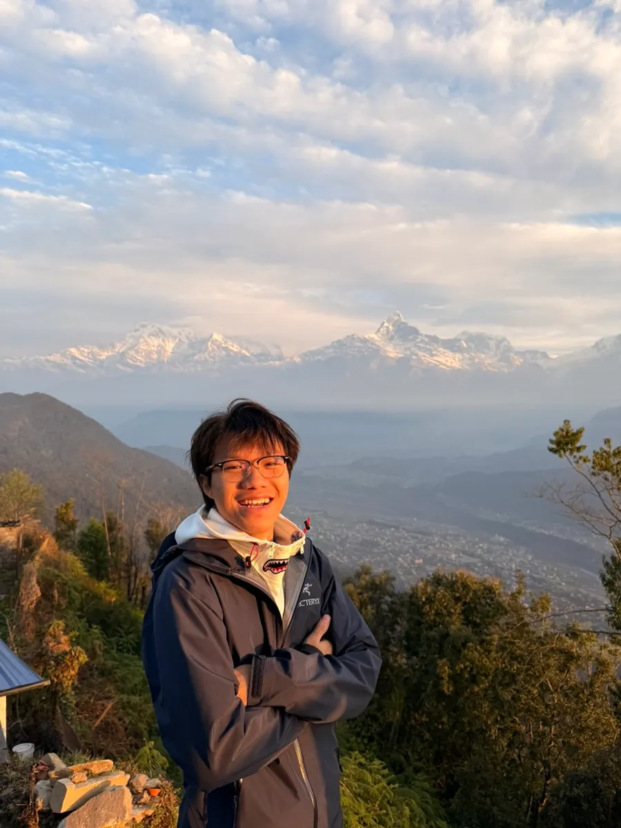 Grinning in a shell jacket at sunrise above Pokhara, with the snow-lit Annapurna range across the valley behind.