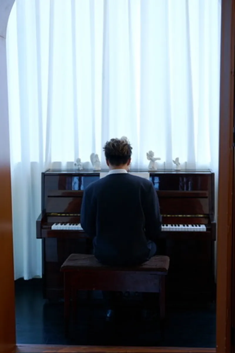 Seen from behind at an upright piano, hands on the keys, a wall of white curtain filling the window beyond.