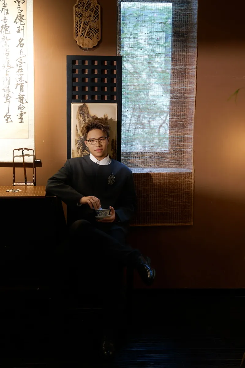Seated in a wood-panelled tea room, holding a small porcelain cup, with a hanging calligraphy scroll and a bamboo blind behind.